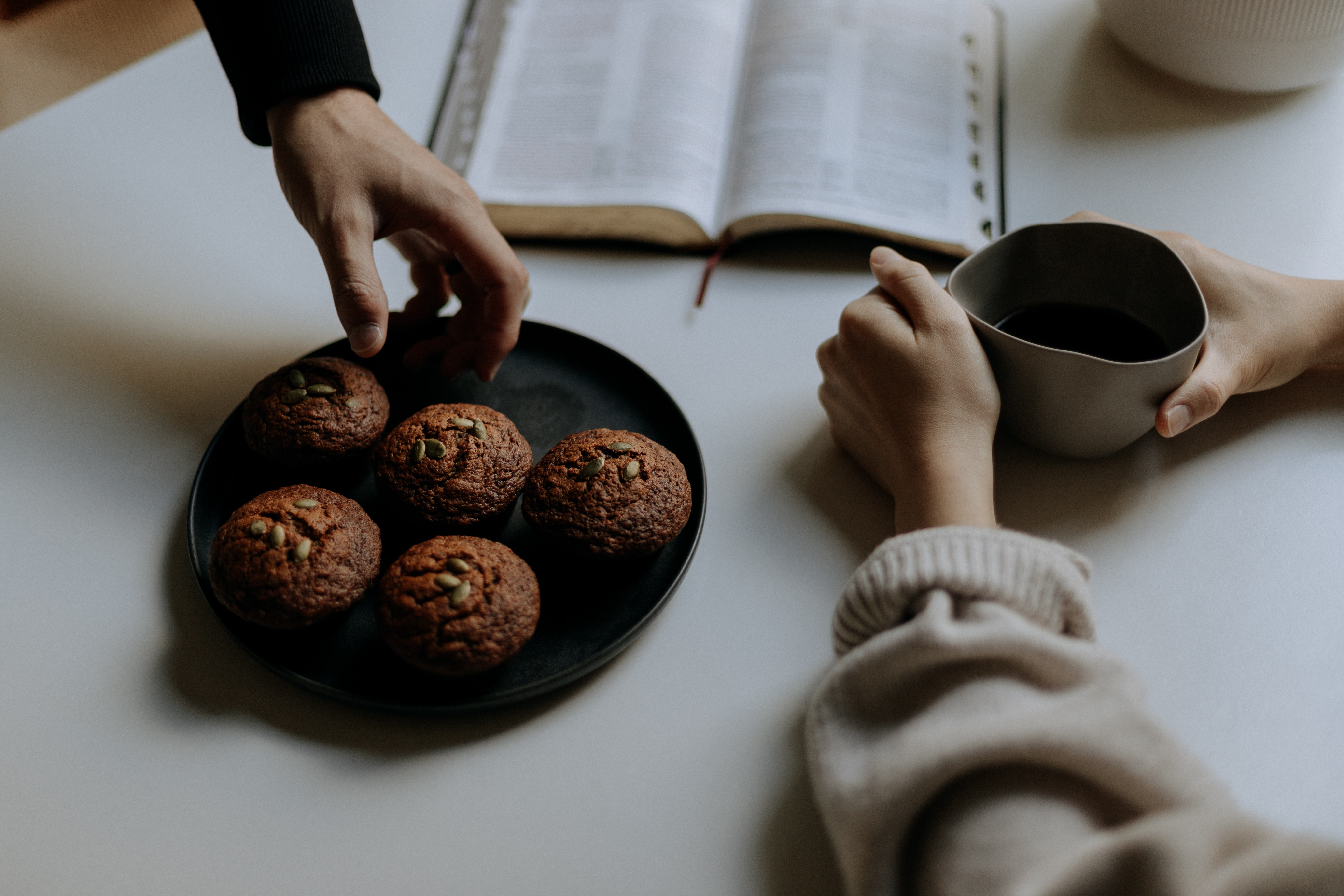 A person grabs a muffin from a plate while another holds a coffee mug. 