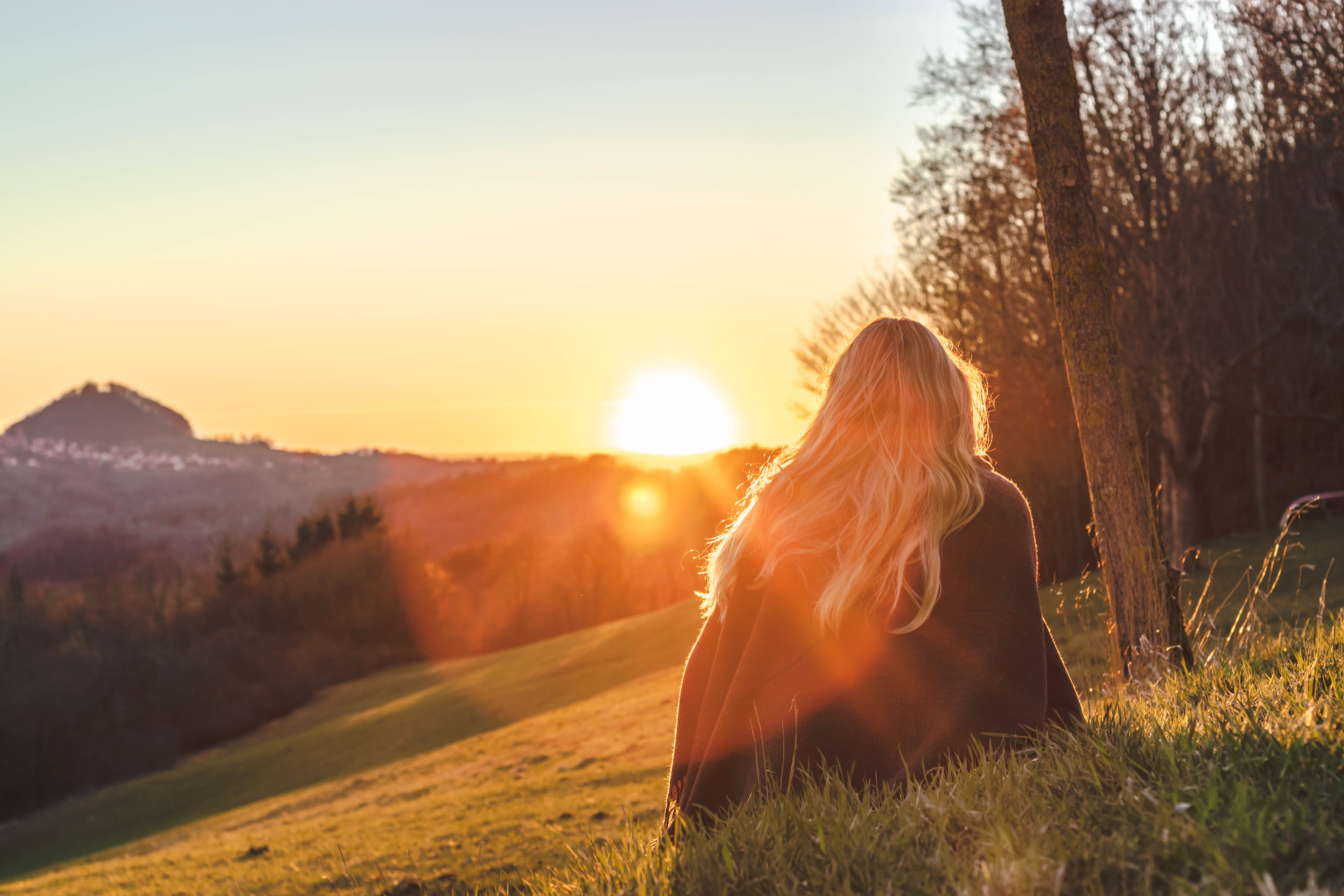 A blonde woman sits in a field watching the sun set. 