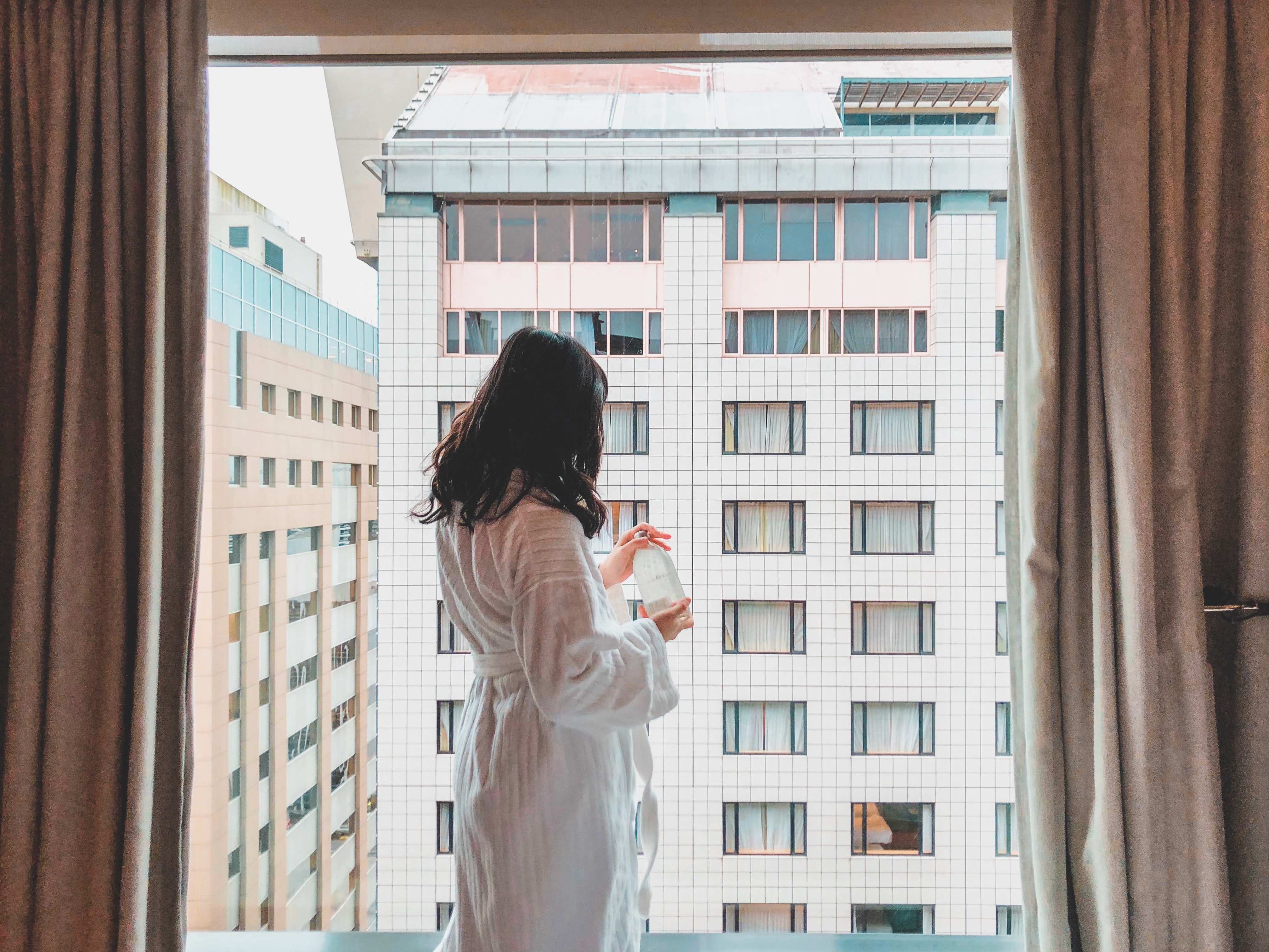 A woman stands in front of a window wearing a white bathrobe.
