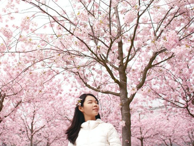 A woman with straight black hair and a white puffy jacket standing among trees with pink blossoms. Her eyes are closed and her expression is very peaceful as a slight breeze appears to blow her hair.
