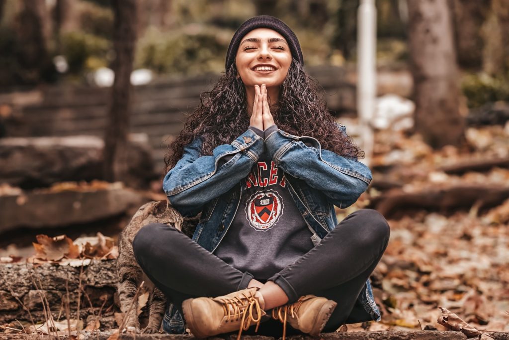 A smiling woman with curly brown hair is sitting on the ground. She's wearing a dark beanie, a gray sweatshirt covered by a blue denim jacket, black jeans and tan laced-up ankle boots. Her legs are crossed and her hands are pressed together as in prayer. The ground is covered in orange autumn leaves and a gray cat with dark stripes stands at her side. The background is blurred, but a few trees can be seen in the distance.