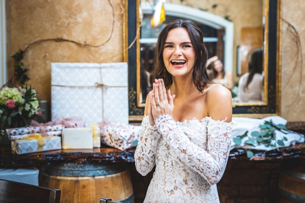 A woman in a white dress stands in front of a gift table at her bridal shower. 