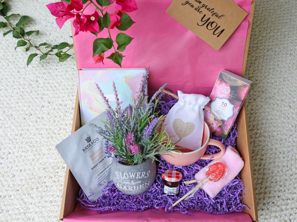 A gift basket holds self care items, such as a facial mask, a journal, a coffee mug, flowers and candy.