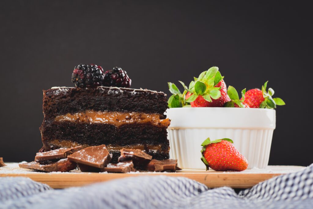 A slice of chocolate cake with blackberries on top sits next to a bowl of strawberries.