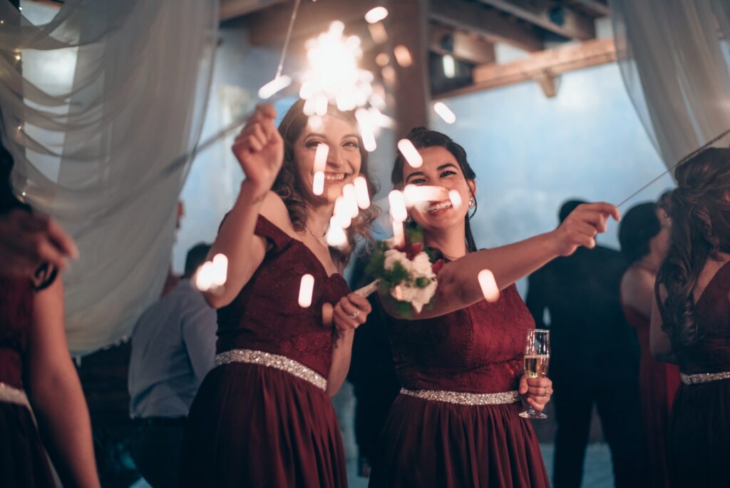 Two bridesmaids in dark red dresses hold sparklers.