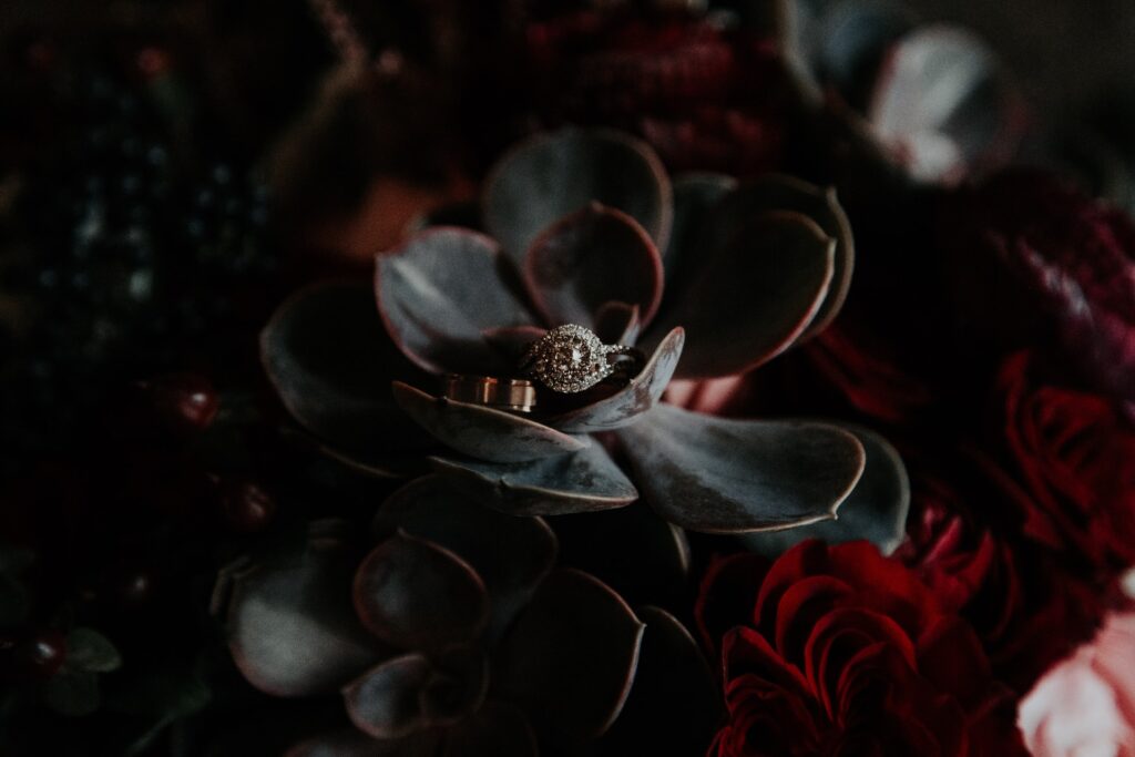 An engagement ring and wedding band sit in a bouquet of dark wedding flowers. 