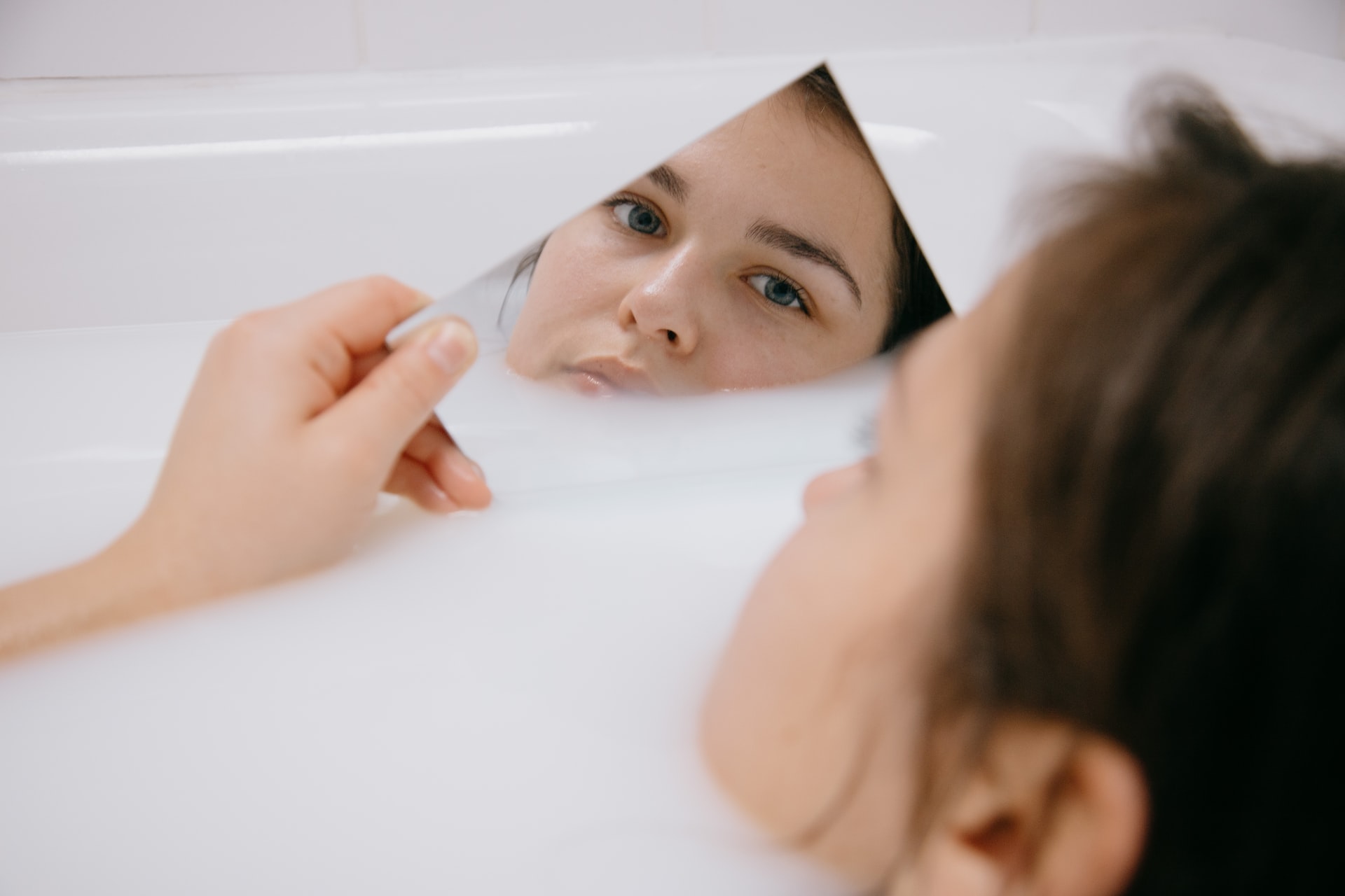 A woman lying in a bathtub looks at her face in a mirror.