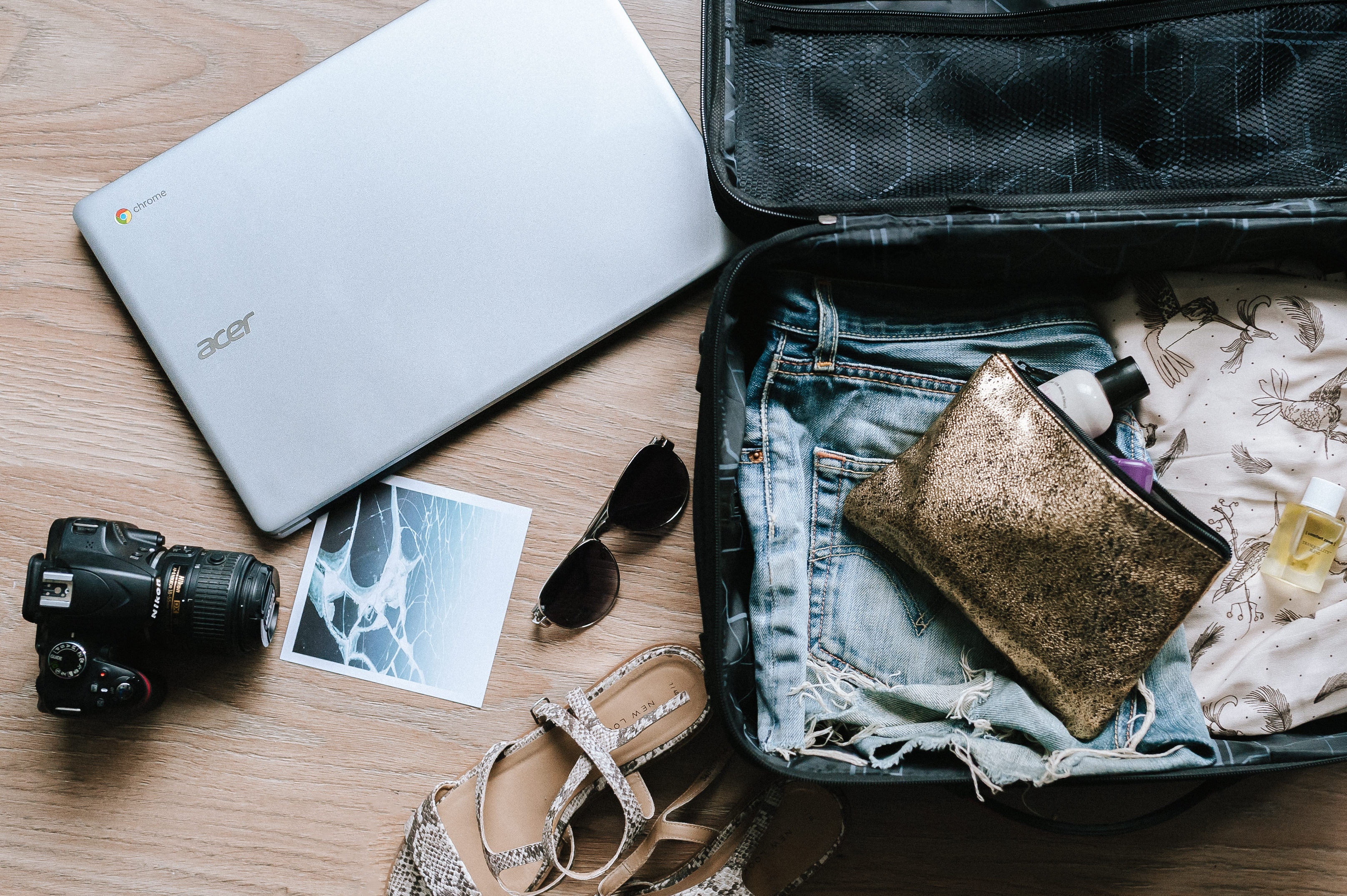 A laptop, camera, sunglasses and clothes sit around an open suitcase.