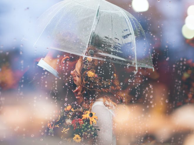 A bride and groom stand under an umbrella in the rain.
