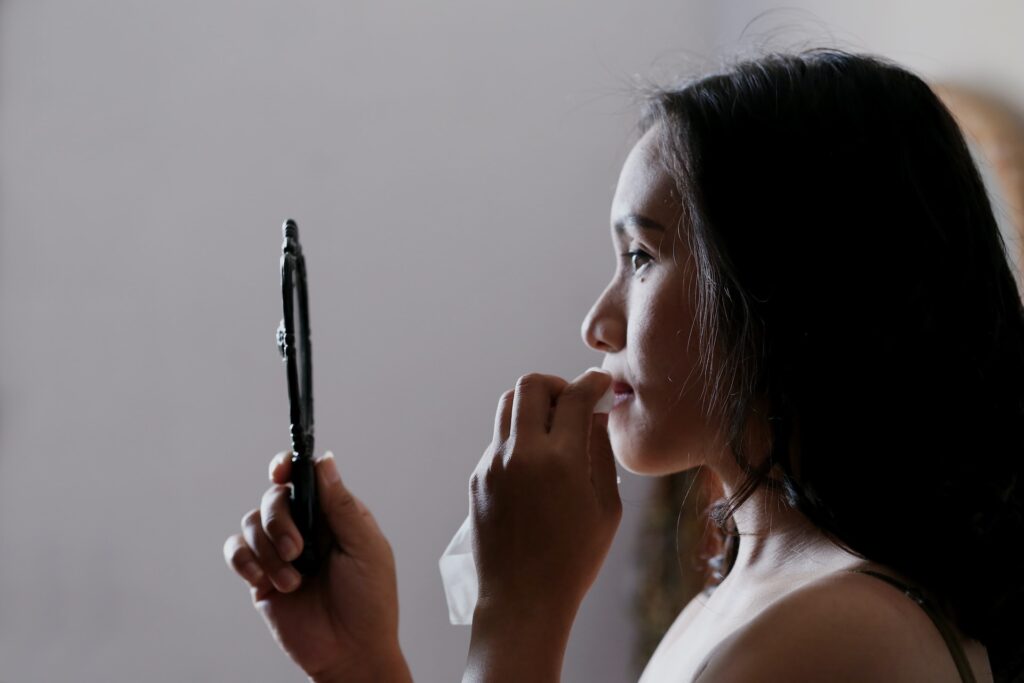 A woman holds a mirror in front of her face and uses a tissue to remove makeup.