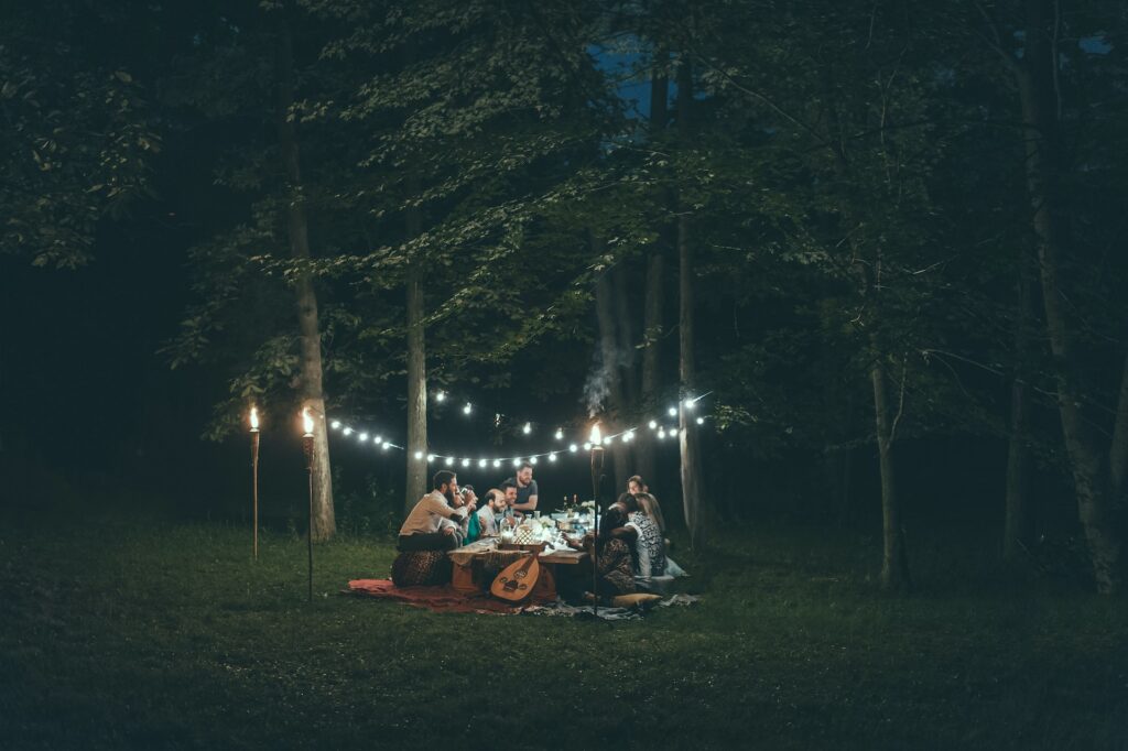A group of people sit at a table in a wooded area with string lights above them.