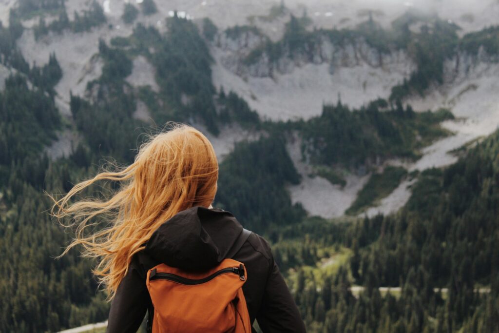 A woman wearing a backpack overlooks a mountain while hiking.