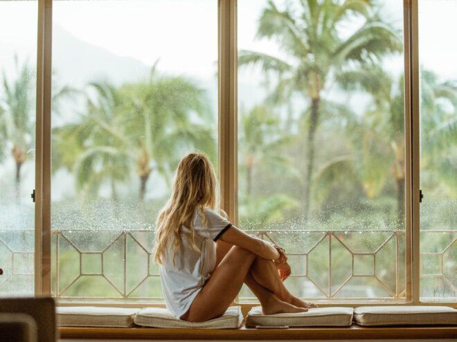 A woman sits in front of a window looking at palm trees.