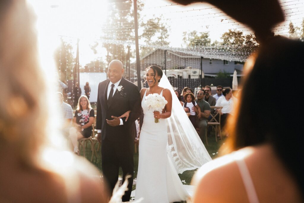 A father walks a bride down the aisle at an outdoor wedding ceremony. 
