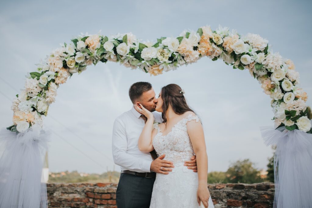 A bride and groom kiss under a floral wedding arch. 