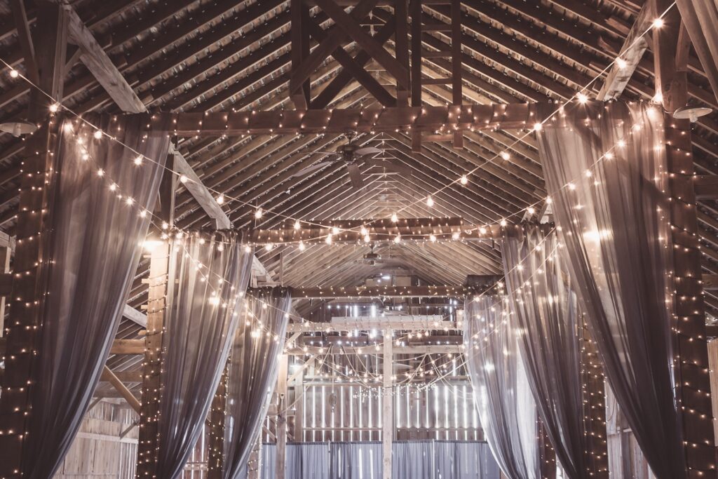 Drapes and string lights hang from the rafters of a barn.