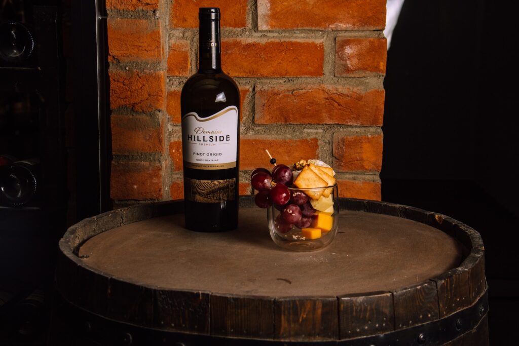 A wine bottle and a cup with cheese and grapes sit on a wine barrel.