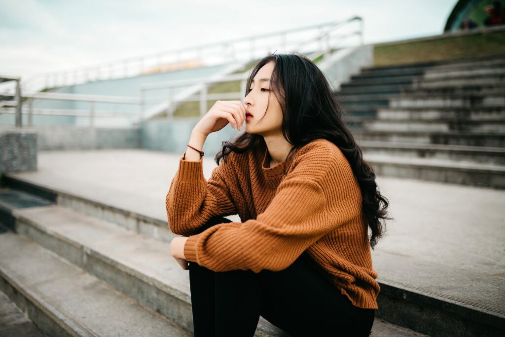 A woman in an orange sweater sits on stairs looking thoughtful.