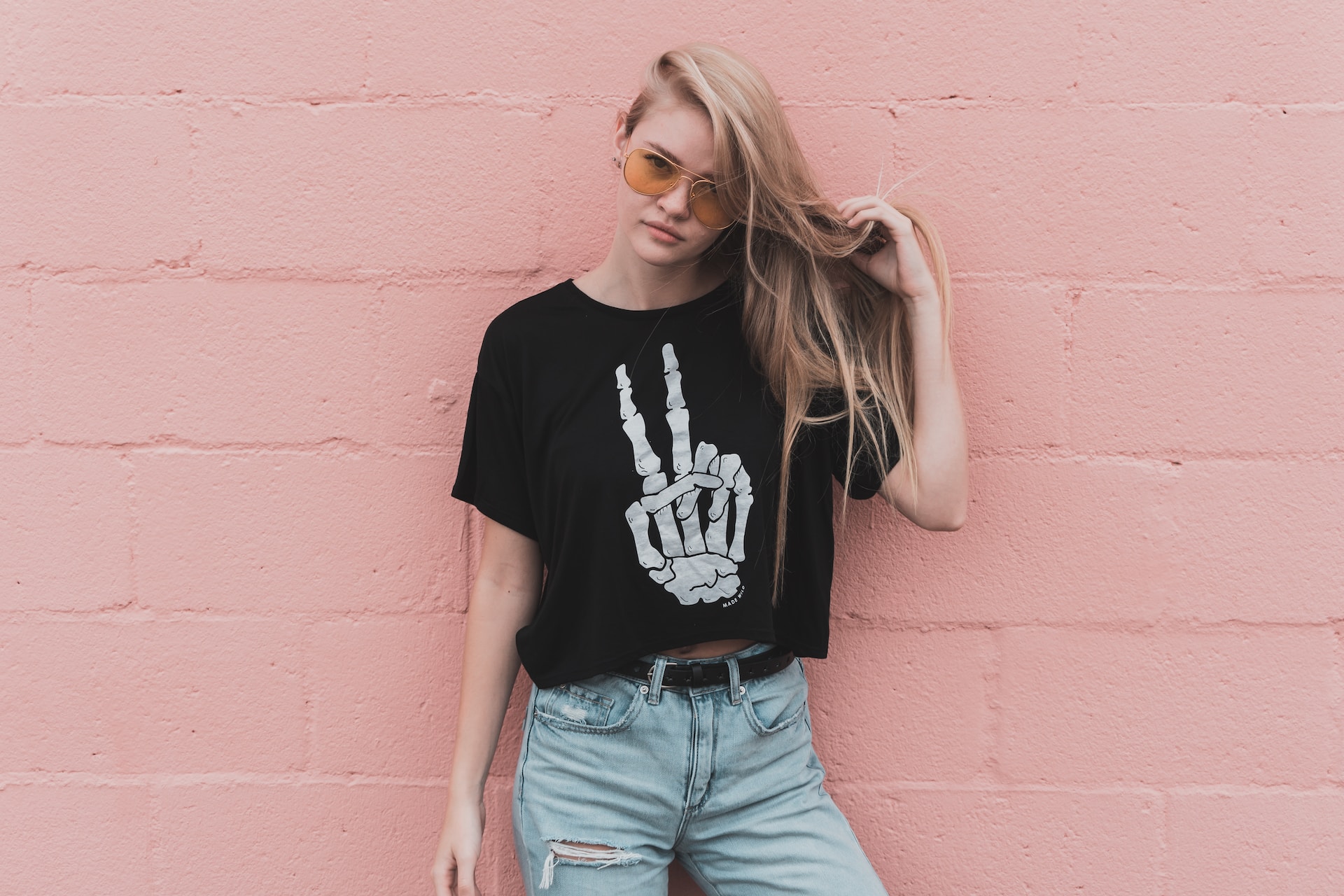 A young woman with long blonde hair wearing a black cropped t shirt with a skeleton hand doing a peace sign standing against a pink brick wall