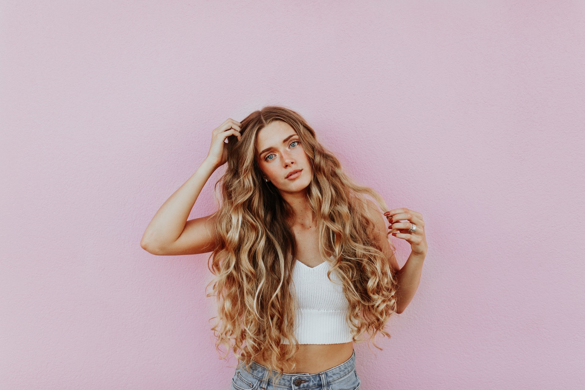 A white woman with long, wavy blonde hair stands against a pink wall