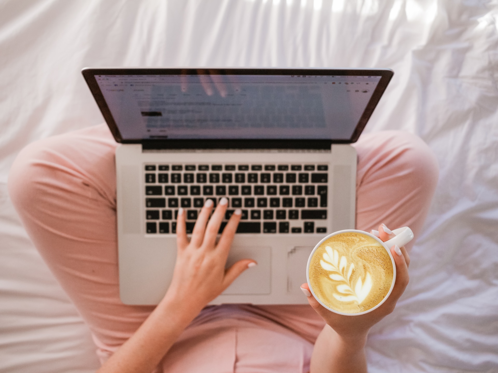 A shot from above of the lower half of a woman wearing pink pants siting on a white bed with a laptop open in her lap and a latte in a mug in her right hand