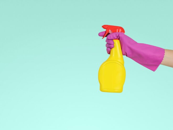 An arm wearing a pink glove holds a yellow and red cleaning spray bottle on a teal backdrop