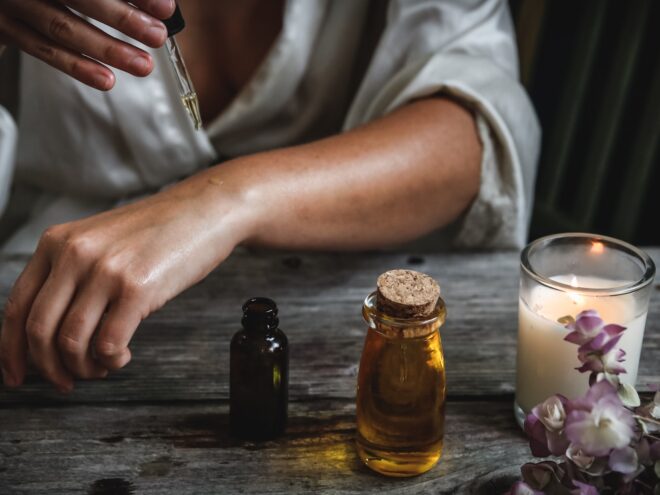 A woman using a dropper to add essential oil to the skin of her left arm sitting at a wooden table with a lit candle and flowers