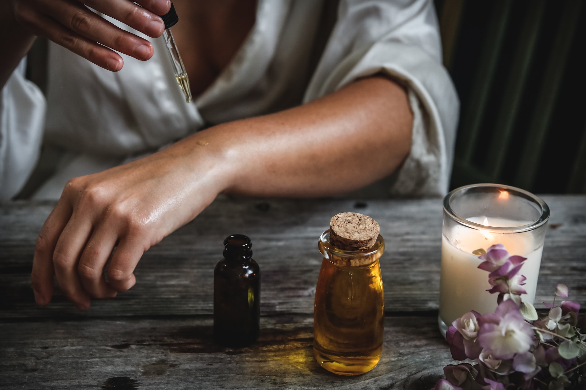 A woman using a dropper to add essential oil to the skin of her left arm sitting at a wooden table with a lit candle and flowers