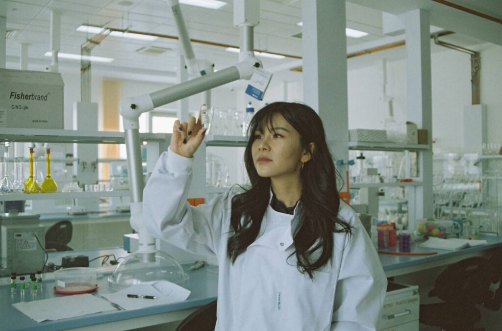 A woman with long black hair and bangs stands in a scientific research lab. The counters behind her are covered in vials and petri dishes. She wears a white lab coat and holds up a glass vial with an unknown liquid in it. She is peering at the liquid.