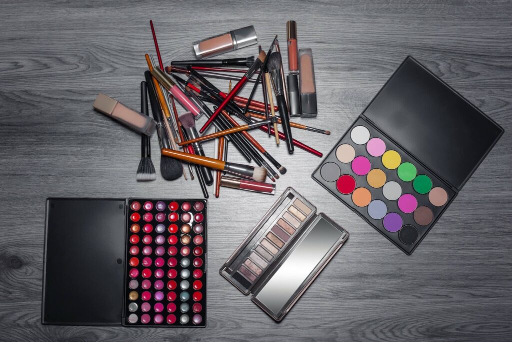 A cluster of makeup products lies on a gray woode surface. The products include two multicolor eyeshadow products, a lipgloss palette, makeup brushes, liquid foundation containers, lip liner and eye liner. None of the products have brands or labels on them.