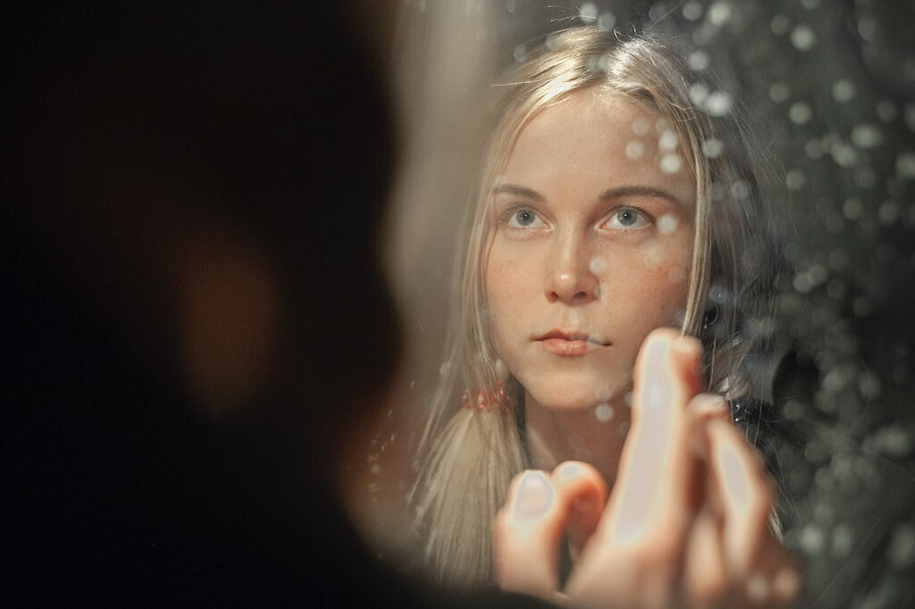 A woman with long blonde hair looks in a mirror. The mirror is covered in water droplets and is slightly foggy. She holds up a hand to her reflection and gazes upward like she's looking for something.