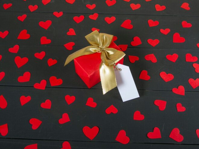 Small gift in red wrapping paper surrounded by red hearts and a gold bow.