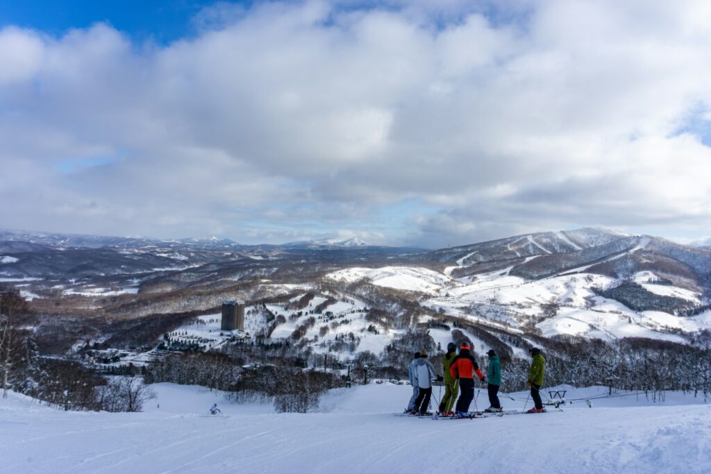 Six skiiers in different colored jackets stand the the top of a slope overlooking a snowy, mountainous region. A brick tower and a small village of buildings with a parking lot sits in the distance at the base of a large hill.