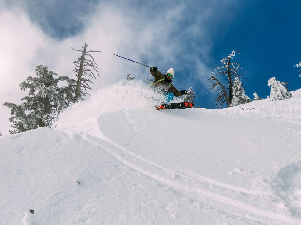 A man makes a sharp turn on his skiis on a snowy slope. Snow covered trees and a blue sky are behind him. Fresh powder flies in his wake.