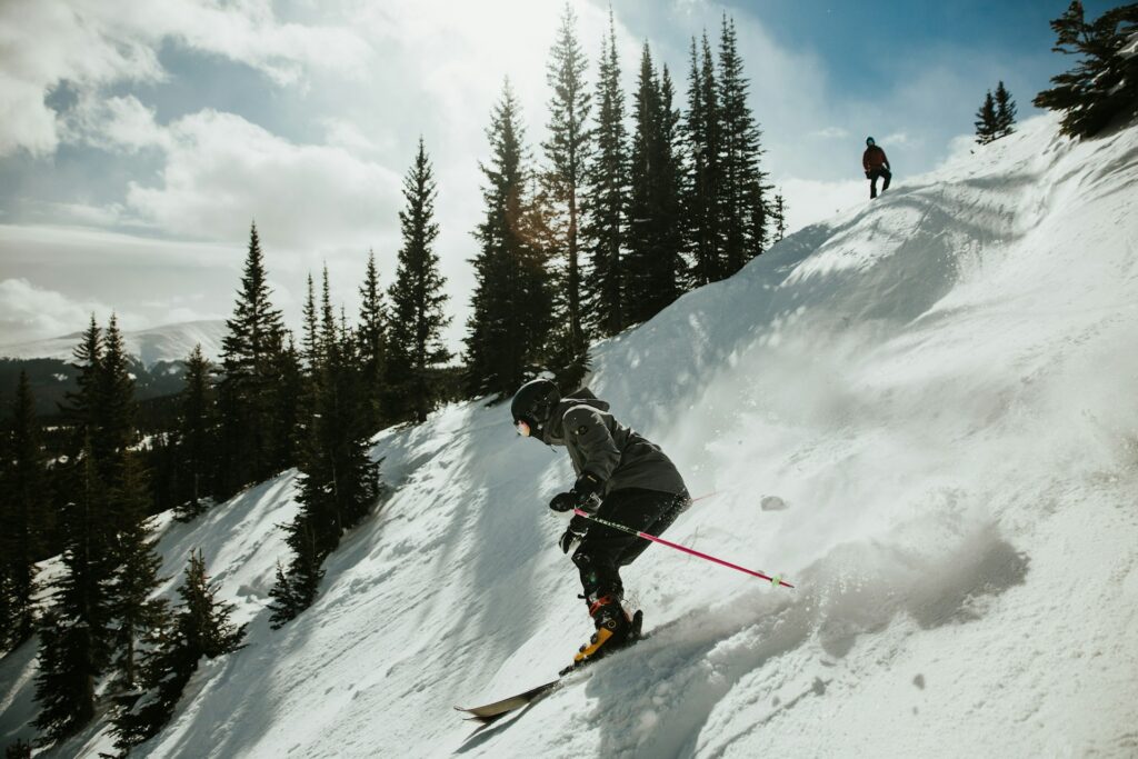 One person watches while another skis down a snowy slopee between feathery green trees under a cloudy blue sky. Both are head-to-toe in ski gear.