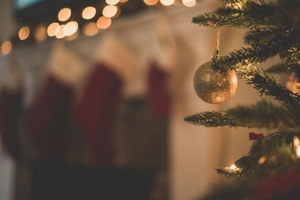 A gold sparkly Christmas wedding ornament hangs from a fir tree branch in front of a blurred fireplace covered in red and white stockings underneath a garland glowing with string lights.