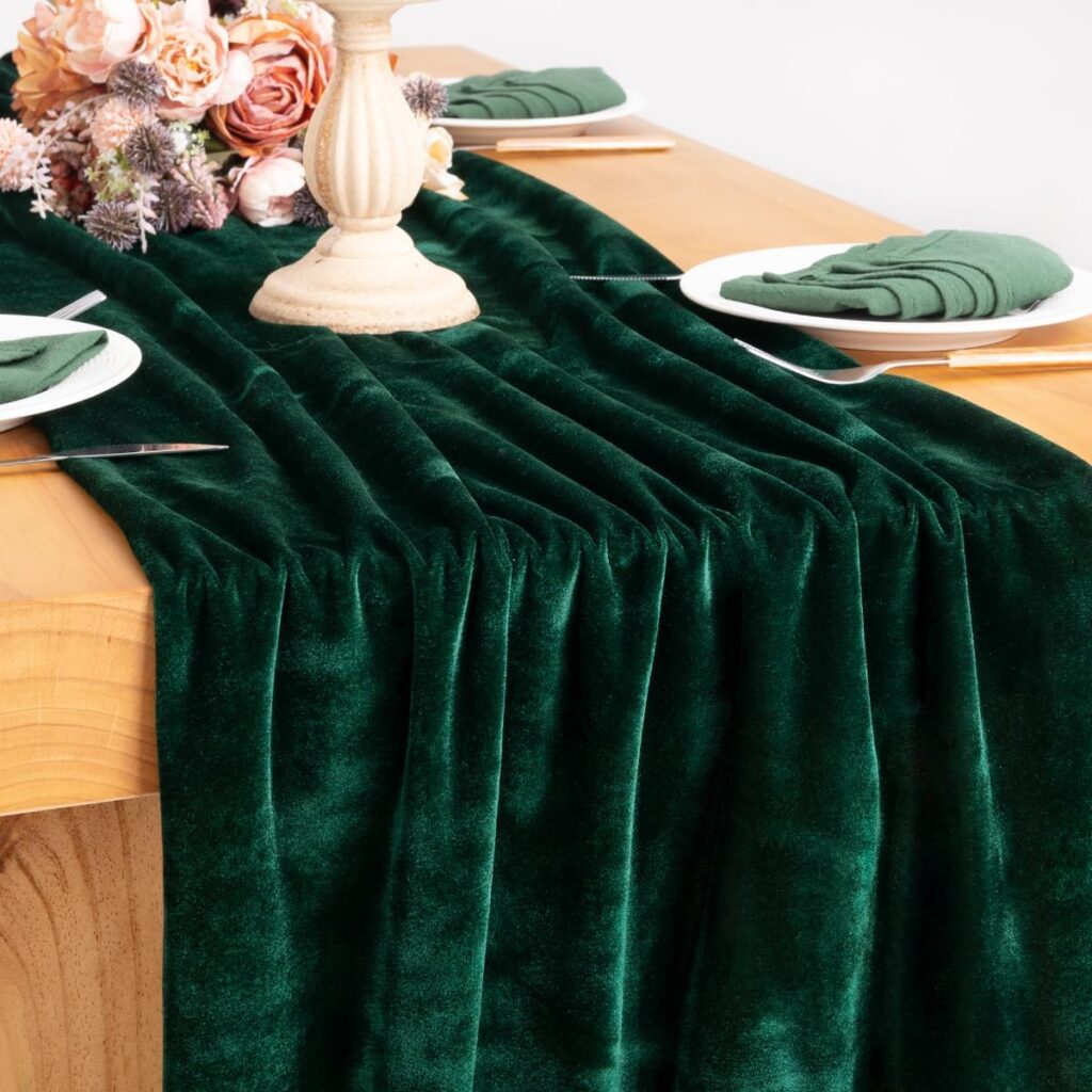 A green velvet table runner is draped over a light brown table. the table is covered in white plates, silverware, green linen napkins, a cream candle holder and a bouquet of cream and pink flowers.