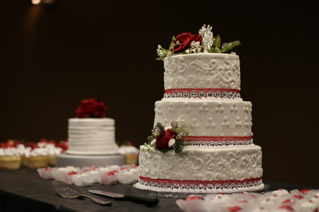 A white wedding cake with three tiers covered in silver jewels, red roses and greener sits on a table covered in a black table cloth. Small cupcakes with white icing and some kind of red decoration on each of them sit around the cake. A smaller white cake with roses on top sits int he background.