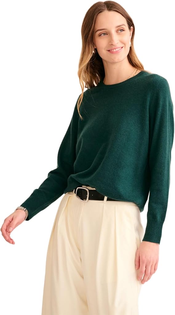 A woman stands in a dark green cashmere sweater, cream pants and a black belt against a white background.