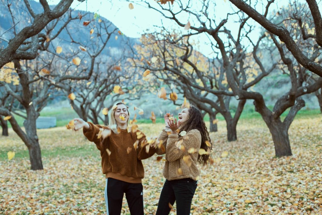 Two women stand in an orchard in the mountains. The trees around them have all lost their leaves. The orange and yellow leaves cover the ground. The women, both wearing brown fuzzy sweatshirts and black pants, toss the leaves in the air above them and laugh.