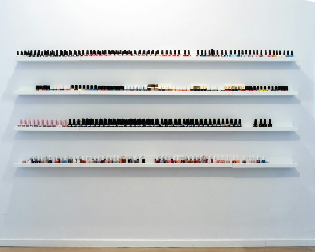 More than 50 nail polish bottles sit on three long white shelves on a white wall, presumably at a nail salon.