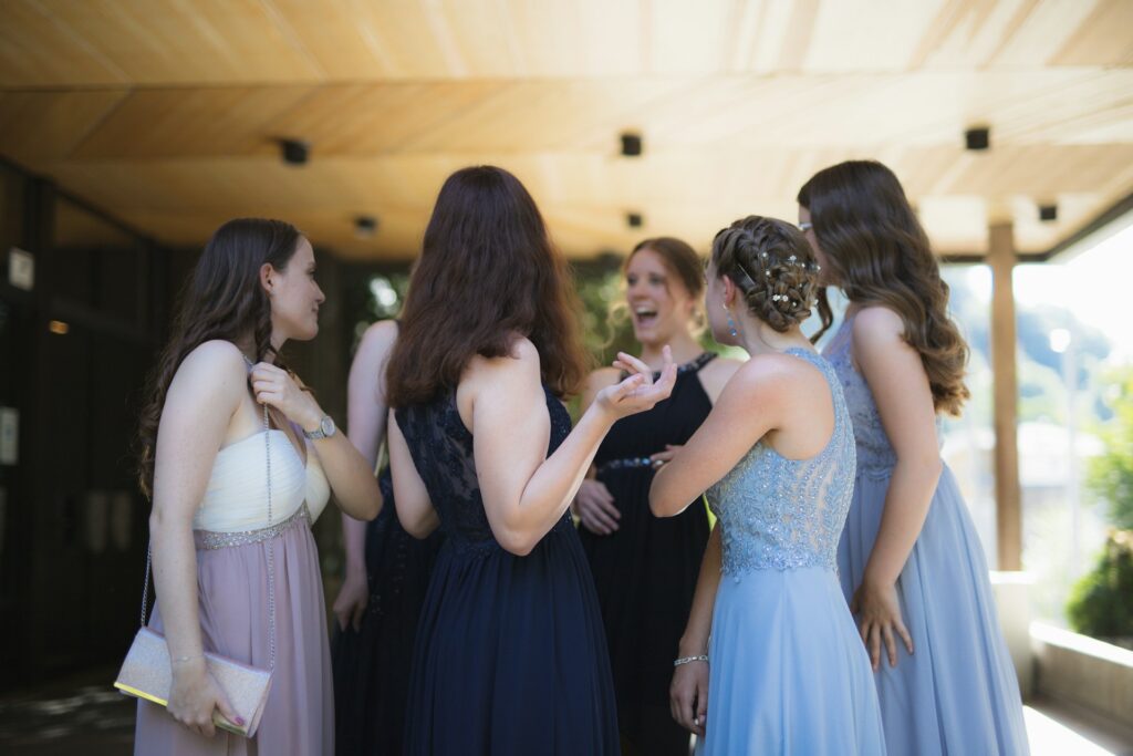 Six women stand around in rehearsal dinner dresses under a wooden awning outside of a building.