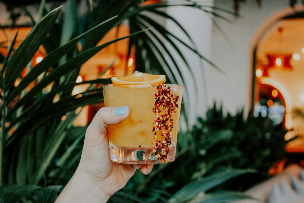 A woman with blue nail polish holds up an apple cider margarita against a background of tall green plants