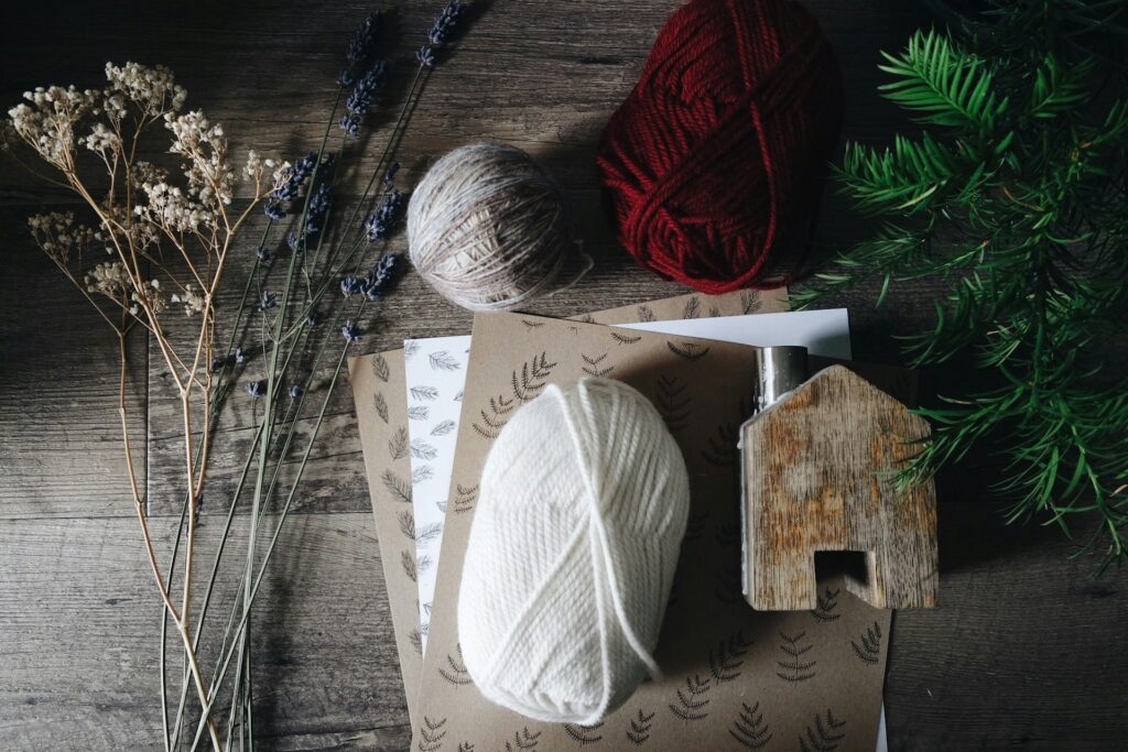 Red, white and gray yarn skeins sit on a gray wooden table with dried flowers, floral brown paper and a faux fir branch. It matches the handmade Christmas aesthetic.