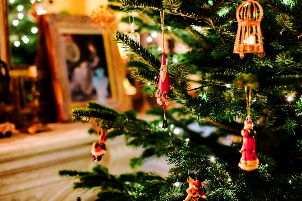 Small wooden ornaments of different Christmas figures hang from a fir tree in a brightly lit room. A white mantle is behind the tree, covered in golden candlesticks, white candles and a golden frame with a picture of people inside it.