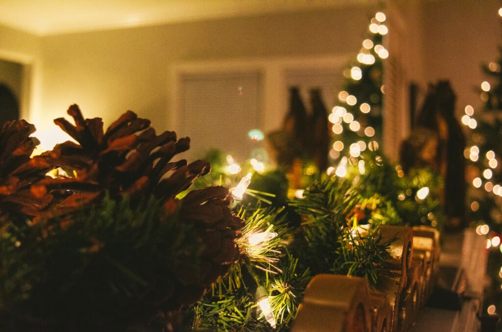 A fir garland with pine cones and string lights sits on a table in a small room. A matching Christmas tree is blurry against some white windows in the background.