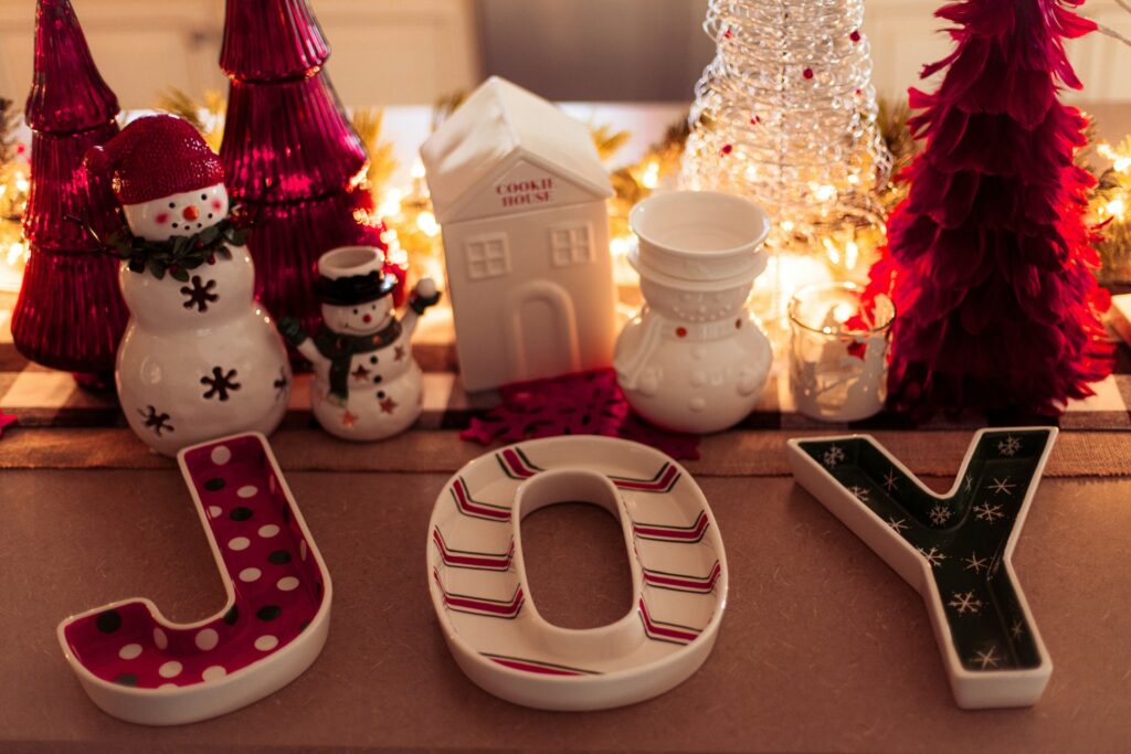 Christmas decor like porcelain snowmen, a cookie house, dip plats spelling out "joy" and a garland lit with string lights behind miniature red Christmas trees decorate a table. They're all on a red and green plaid table runner.