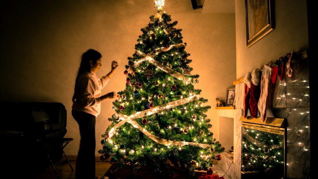 A woman in a white sweatshirt and black pants decorates a brightly lit Christmas tree. It's covered in godlen ribbon and multi-colored glass ornaments. The lights are off in the room, so the light from the tree illuminates a marble fireplace covered in red stockings and pictures in golden frames on the walls.