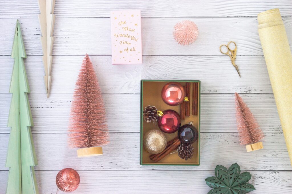 Mini pink Christmas trees, wooden trees, golden scissors and ornaments in shades of pink, silver and black sit on a white wooden table. They're a great starting point for a pink Christmas aesthetic.