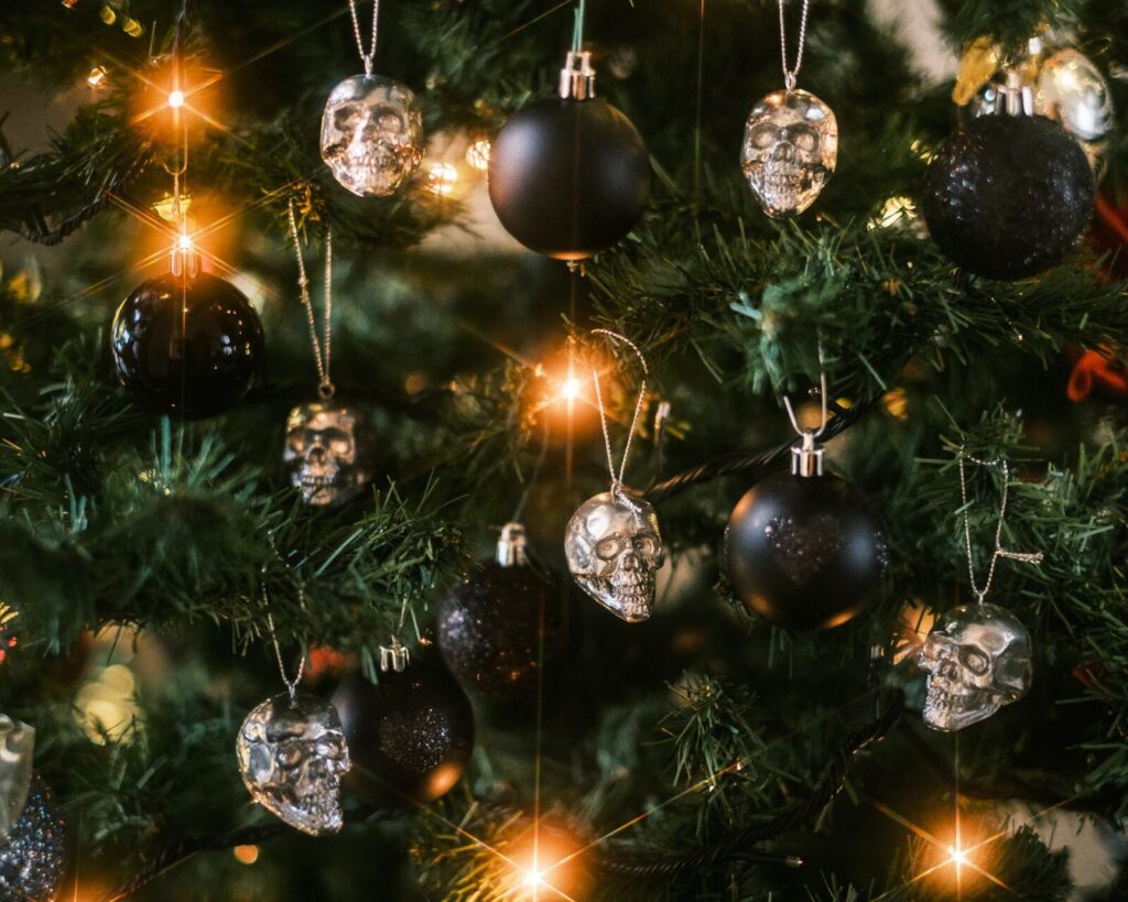 Black ornaments covered in sparkles and silver skull ornaments hang from a fake fir Christmas tree among yellow string lights. They inspire people to try a dark Christmas aesthetic.